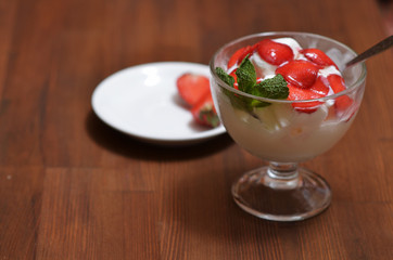 Vanilla ice cream with sliced strawberries and mint leaves in a glass bowl with a spoon on a wooden table