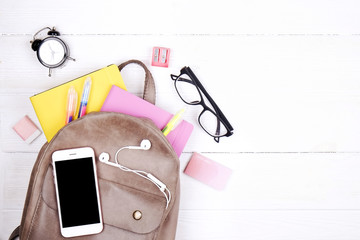 Hipster grey leather backpack, full of school supplies, blank screen cell phone, earphones, pink & yellow notebook, glasses, mini alarm clock. Back to school concept. Close up, copy space, flat lay.