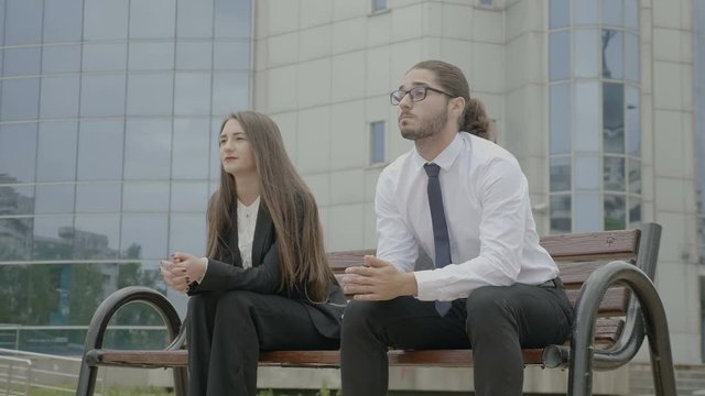 Young beautiful businesswoman and businessman sitting on a bench in front the corporation looking in the eyes bumping fists and dancing
