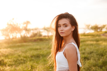 Photo of brunette young woman with long hair wearing white dress, looking at you while walking in nature during bright sunny day