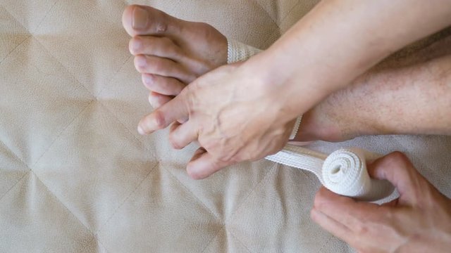 First aid man applying bandage for injured foot and ankle sitting on couch at home. Health care damaged sprained foot problems