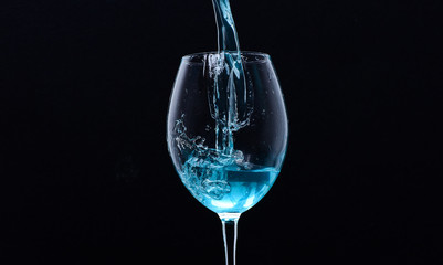 Wineglass filling with water with splashes on black background. Refreshing drink concept. Cocktail with blue liquid in glass. Glass with blue water pouring with liquid with splashes and drops