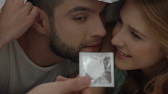 Hand offering condom to happy couple lying in bed, safe sex, intimate relations