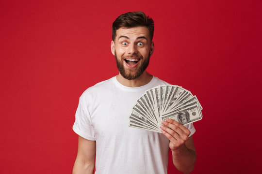 Photo of happy european man in casual t-shirt smiling and holding fan of money in dollar banknotes, isolated over red wall