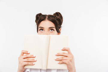 Photo of dreaming teenage girl with double buns hairstyle reading and looking upward while covering face with book, isolated over white background