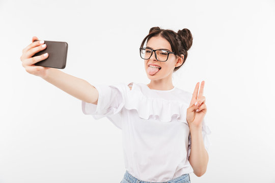 Sly cheerful woman with double buns hairstyle wearing eyeglasses fooling around and showing peace sign while taking selfie at smartphone, isolated over white background