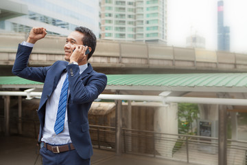 Businessman raising his fist  in the air during callling the phone, with office building background - business success, achievement, and win concepts
