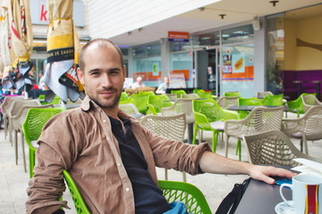 Adult man with casual clothing sitting at a table outside a coffee shop smiling and looking and the camera