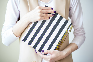 Close-up photo of manicured woman's hands hold an elegant notebook with black and white strips. Purple nail polish