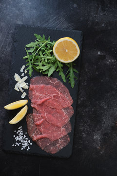 Above view of a stone slate tray with carpaccio made of raw marbled beef, arugula, lemon and parmesan cheese over dark scratched metal background