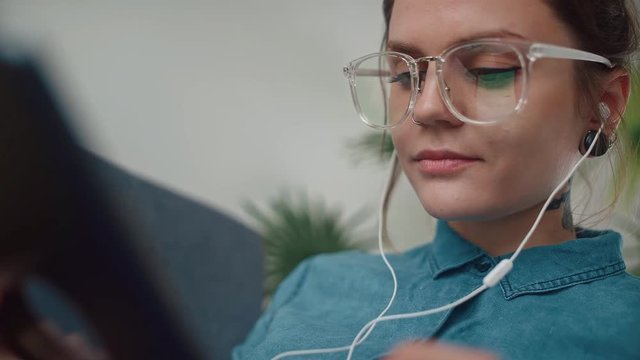 Young modern girl with glasses and headphones listens to music, smiles easily and looks at tablet screen. Concept internet, virtual communication, work with tablet. Stylish young girl with a gadget.