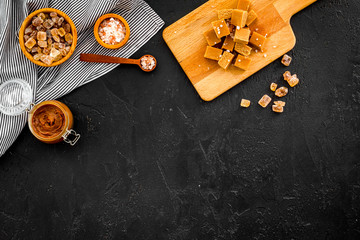 Salted caramel background. Melted caramel in glass jar, caramel cubes on cutting board, sugar and salt on black desk top view copy space