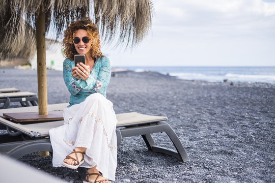 nice smile beautiful middle age lonely woman enjoy the beach sitting on a seat with ocean in background. using smartphone to connect with friends at home or to work with a team online business.