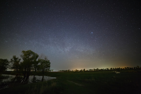 Space with the stars of the Milky Way in the night sky. The landscape with the river and trees is photographed on a long exposure.