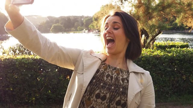 Happy woman taking selfie with smartphone in park in front of lake smiling