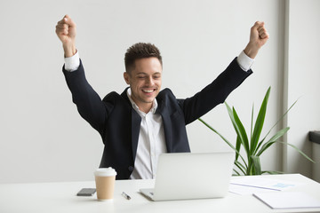 Happy millennial businessman raising hands feeling joy celebrating success or good online result, win on stock looking at laptop, man in suit excited by great email news or deal enjoying victory