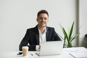 Smiling millennial businessman in suit sitting at work desk with laptop, happy professional looking...