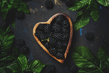 Fresh blackberries with leaves in wooden bowl in the shape of  heart, low key, top view