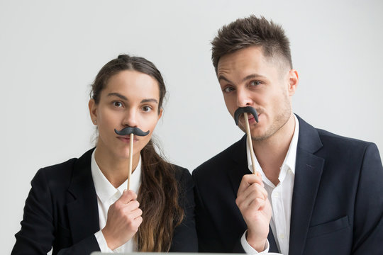 Funny male and female colleagues in suits holding fake mustache, businessman and businesswoman making silly faces, millennial coworkers grimacing looking at camera, creative team head shot portrait - Powered by Adobe