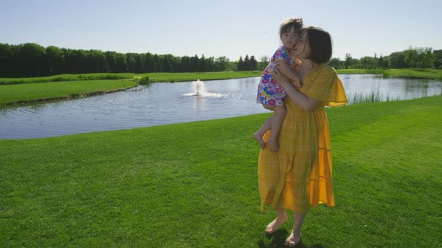 Young mother in yellow dress walking barefoot on grass near pond embracing and carrying little toddler daughter with down syndrome in arms. Sunny girl and mother enjoying summer outdoors. Steadicam