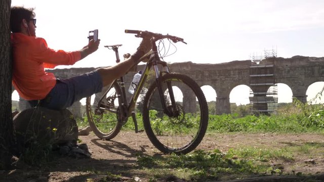Cyclist with bike sitting on a rock under a tree taking selfies in front of ancient roman aqueduct. Young attractive athletic man with orange sportswear and backpack in parco degli acquedotti in Rome