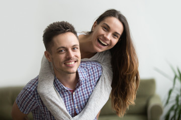 Obraz na płótnie Canvas Smiling husband piggybacks cheerful wife at home, millennial couple in love embracing looking at camera, young real estate owners having fun in new house, happy man and woman dating headshot portrait