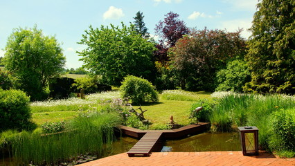 Fototapeta na wymiar malerischer naturbelassener Garten mit Teich an roter Terrasse mit Holzbrücke und Blumeninseln