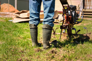 Farmer in rubber boots and blue jeans plows a soil with smal motor tractor.