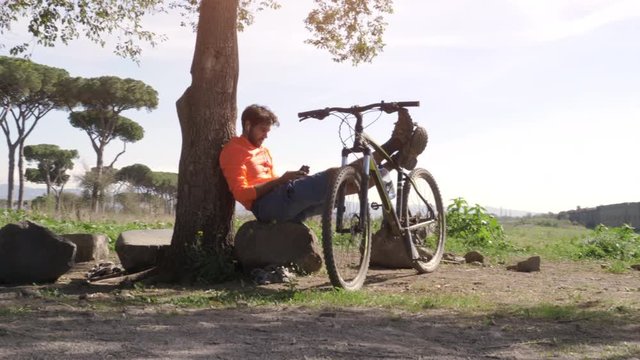 Cyclist with bike sitting on a rock under a tree taking selfies in front of ancient roman aqueduct. Young attractive athletic man with orange sportswear and backpack in parco degli acquedotti in Rome