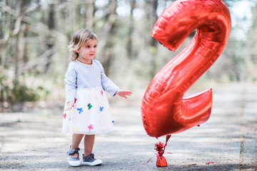 Blonde white female toddler wearing a grey and white dress with multi colored butterflies holding and looking at a red number two balloon.