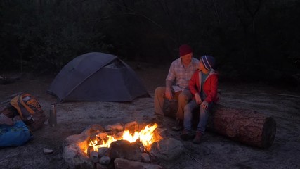 Father and son sitting by a camp fire in the evening on a camping trip together in nature - Powered by Adobe