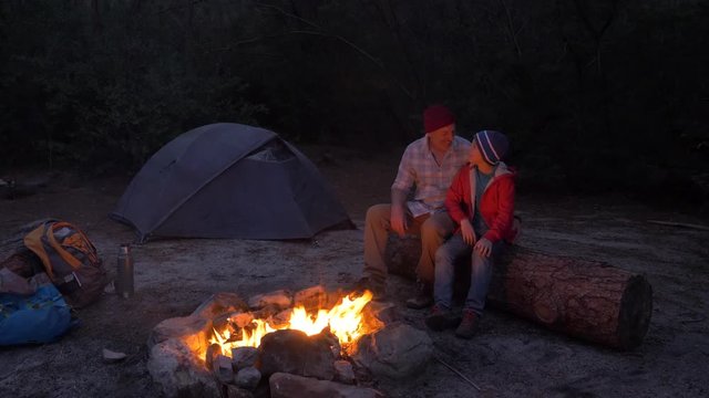 Father and son sitting by a camp fire in the evening on a camping trip together in nature