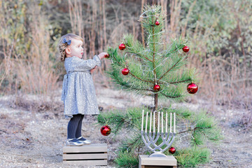 Blonde white toddler girl standing on a wood box wearing a grey dress and grey leggings decorating a tree with red decorations and a menorah.