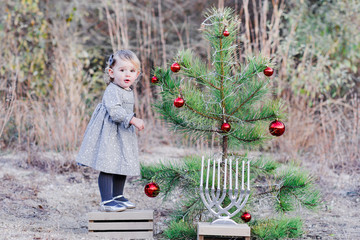 Blonde white toddler girl standing on a wood box wearing a grey dress and grey leggings looking at the camera next to a tree with red decorations and a menorah.