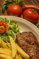 Close up of detailed well done grilled New York steak with french fried potatoes, tomatoes, sausage. served in white plate on cutting board on wooden table background