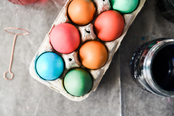 Top close up view of multi-colored eggs drying in a cardboard egg container on top of parchment paper surrounded by dyed water in mason jars..