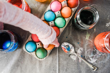 Top view of toddler hand placing a dyed orange egg in a cardboard container with multi-colored eggs drying on top of parchment paper surrounded by dyed water in mason jars and utensils.