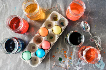 Top view of multi-colored eggs drying in a cardboard egg container on top of parchment paper surrounded by dyed water in mason jars..