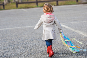 Backside view of a blonde female toddler wearing a khaki rain coat with red boots holding a rainbow ribbon toy running away from camera.