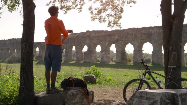 Cyclist with bike standing on a rock under a tree looking at ancient roman aqueduct. Young attractive athletic man with orange sportswear and backpack in parco degli acquedotti in Rome