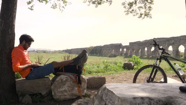 Cyclist with bike sitting on a rock under a tree writing on laptop in front of ancient roman aqueduct. Young attractive athletic man with orange sportswear and backpack in parco degli acquedotti in
