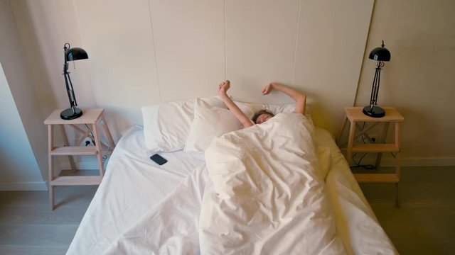 Young Lady is Waking Up in a Cozy Bedroom and Turning Off the Alarm Clock on her Phone in the Sunny Morning