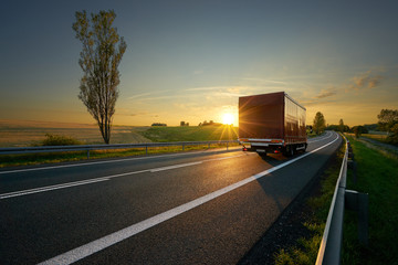 Red small truck driving on asphalt road around farm fields in rural landscape at sunset