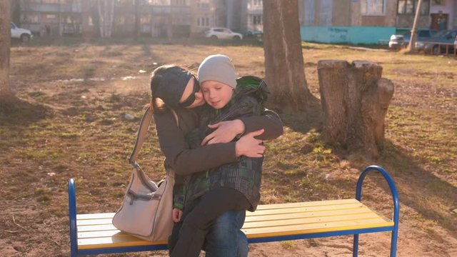 Mom kisses and hugs her son sitting on a bench in the courtyard of a multi-storey building.