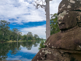 A statue on the south gate at Angkor Thom. View of the Clondy sky and the tree reflection on the river.