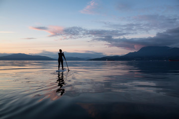 Adventurous girl on a paddle board is paddeling during a bright and vibrant sunset. Taken near...