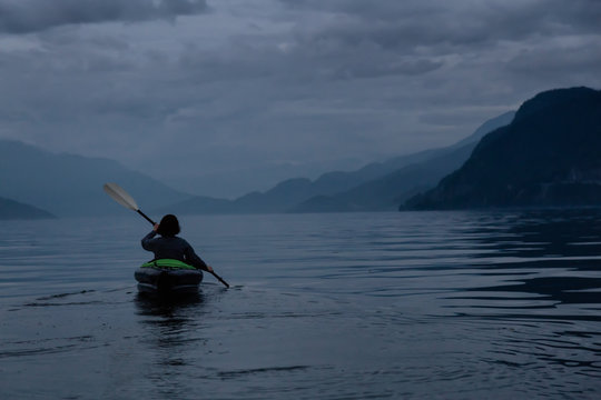 Adventurous woman kayaking during a dark gloomy evening surrounded by Canadian Mountain Landscape. Taken in Howe Sound, North of Vancouver, BC, Canada.