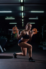Fototapeta na wymiar A strong, muscular, girl in black shorts, performs a squat with a barbell and a leg step. Dark gym.