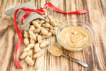 The bag of raw peanuts in shell, glass bowl of peanut butter on wooden background with copy space. Flatlay. Heathy food.