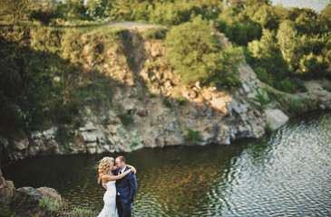 Beautiful and lovely newlyweds are hugging cute on the cliff, against the background of rocks and river. Wedding portrait of a stylish bride and a cute bride. Smiling newlyweds.